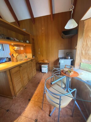 A kitchen with wooden cabinets, a dining area with metal chairs, a small table, and a kettle on the stove. - Cabañas Bahía Celeste (Puerto Varas)