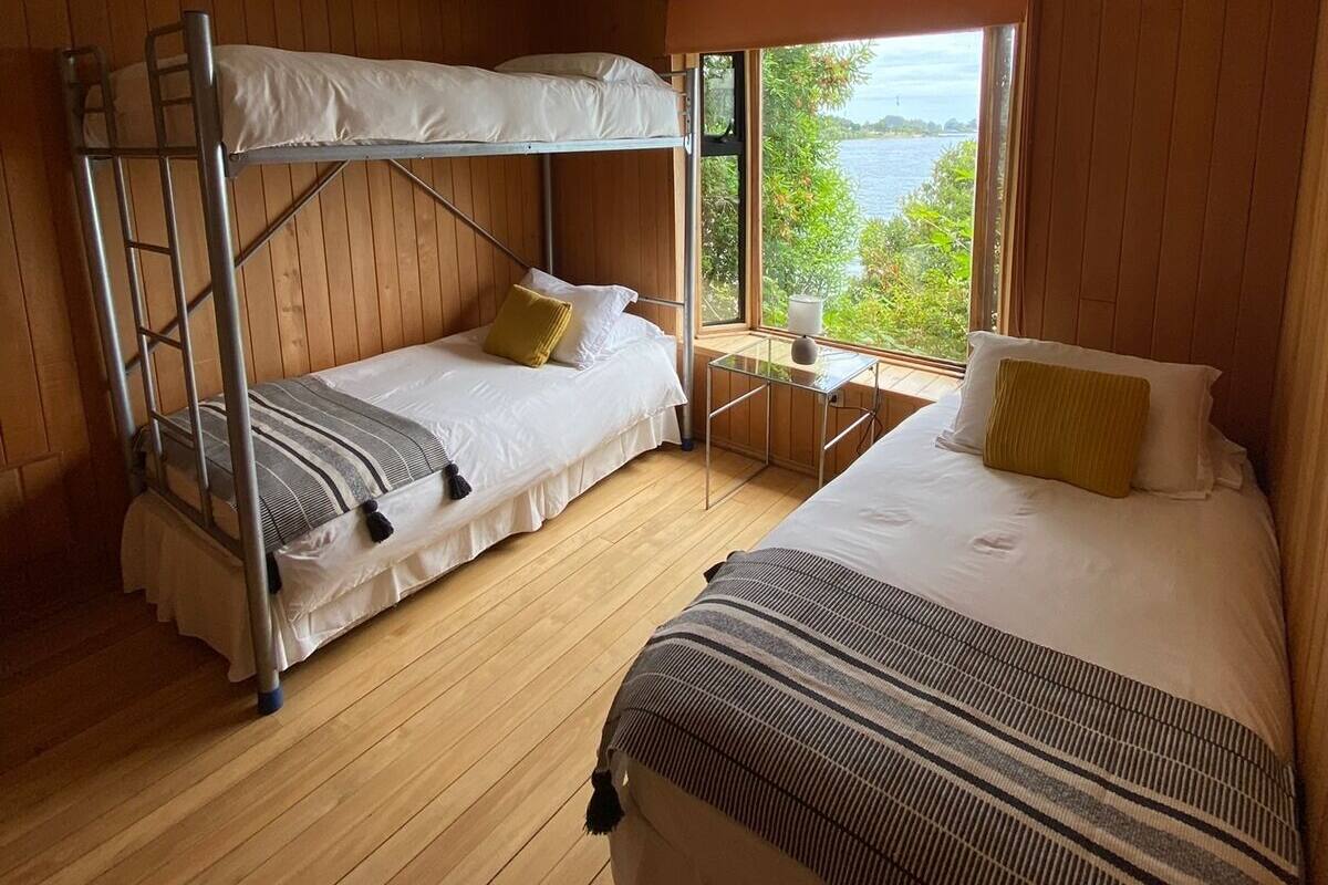 Habitación con dos camas, una litera y una individual, piso de madera, ventana con vistas y una pequeña mesa con lámpara.