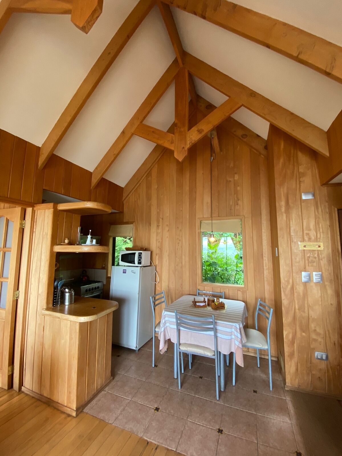 A wooden cabin with a kitchen area, a dining table with chairs, and a window with a view of greenery.