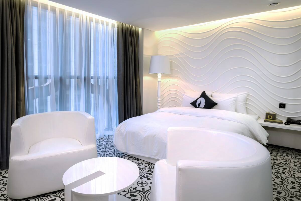 A modern hotel room with a large bed, two white armchairs, a small round table, and a decorative wall.