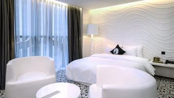 A modern hotel room with a large bed, two white armchairs, a small round table, and a decorative wall.