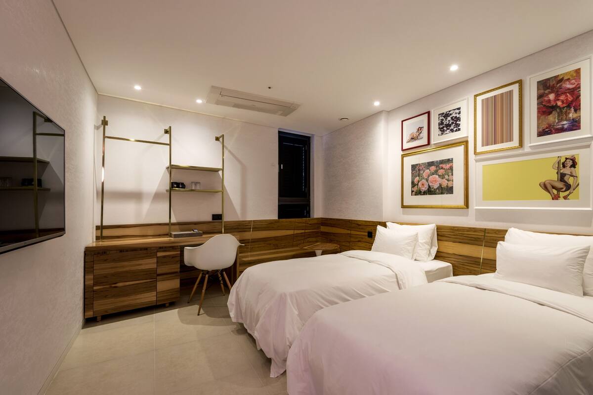 A modern hotel room with two beds, a desk, a chair, and artwork on the walls.