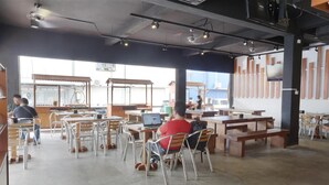 Um café amplo com pé-direito alto, cadeiras de metal e mesas de madeira.
