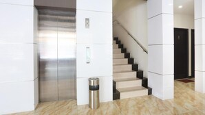 Um elevador moderno, uma escadaria e uma lixeira numa área de lobby limpa e bem iluminada.