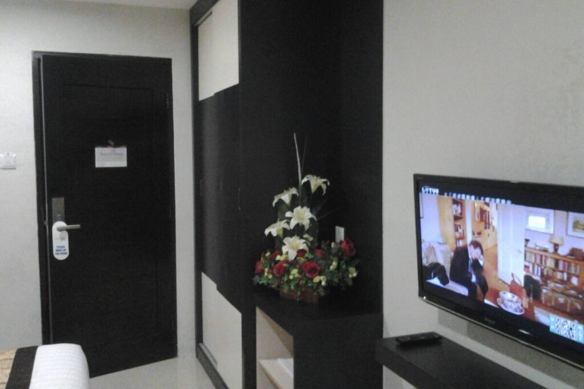A room with a black door, a television displaying a show, and a flower arrangement on a shelf.