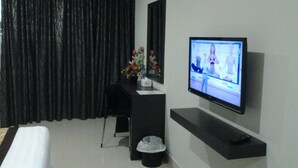 Quarto de hotel com televisão de tela plana na parede, uma escrivaninha com um vaso de flores, uma lixeira preta e uma cadeira preta.