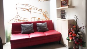 A red sofa with two grey cushions, a wall with a ship illustration, a small shelf with a picture frame, and a vase with flowers. - Evora Hotel (Surabaya)