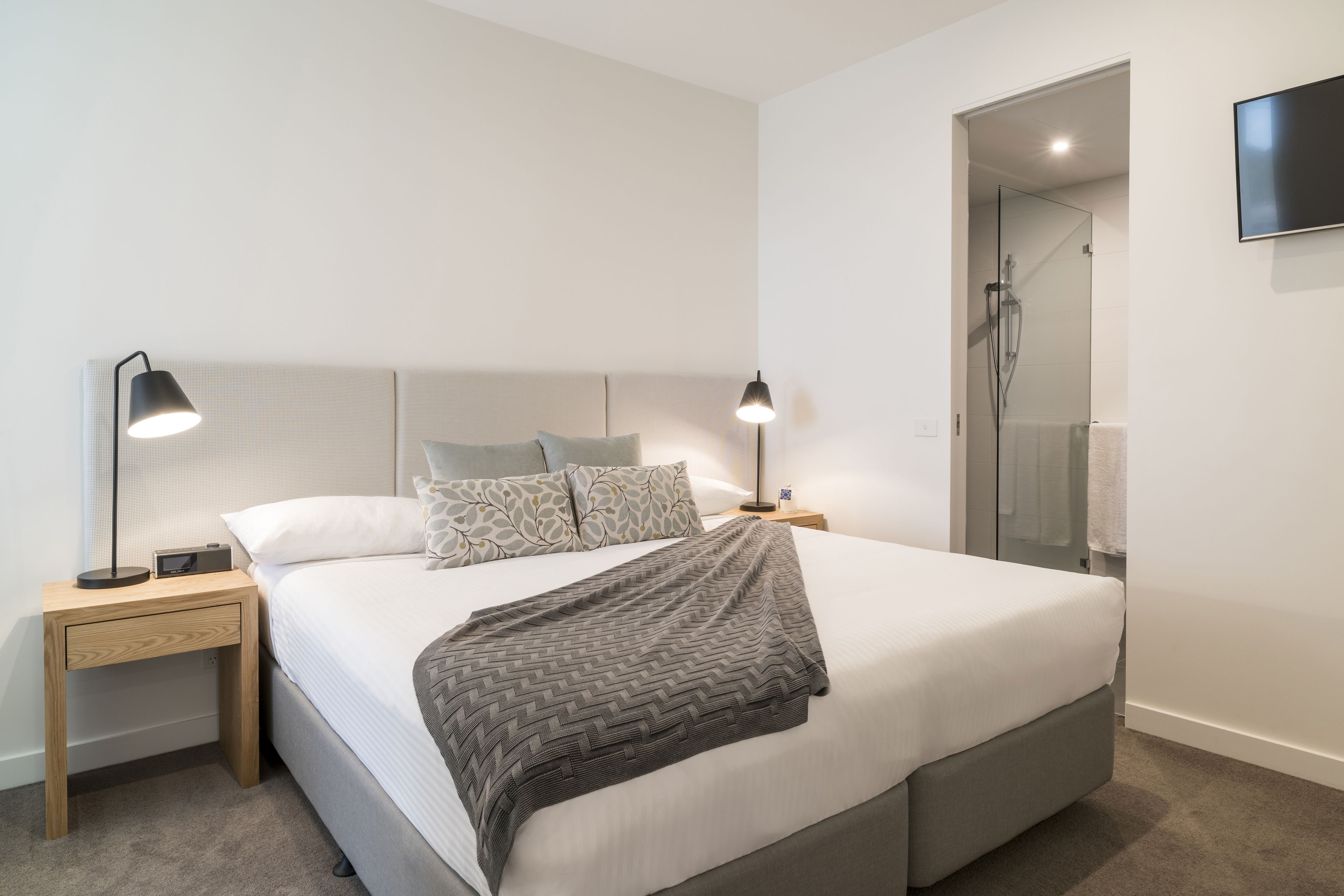 A modern bedroom with a large bed, two bedside tables with lamps, a glass shower door, and a mounted TV.