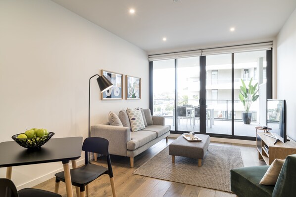 A modern living room with a sofa, a coffee table, a dining table with a bowl of fruit, and a large window with a view of the outside. - Caroline Serviced Apartments Sandringham (Sandringham)