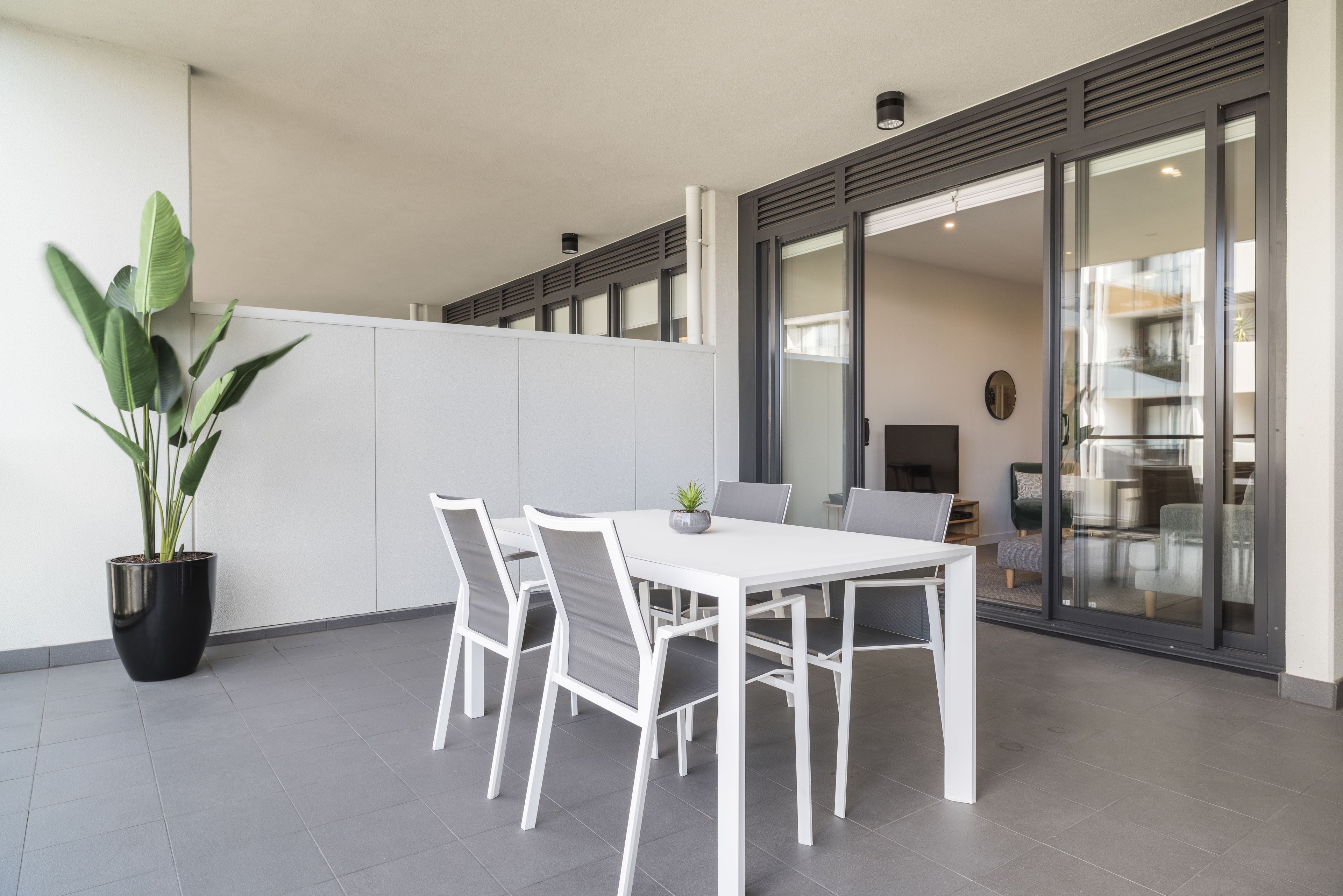 Uma área moderna para refeições ao ar livre com uma mesa e cadeiras brancas, um vaso de plantas e uma porta de vidro deslizante que dá para um espaço interno.