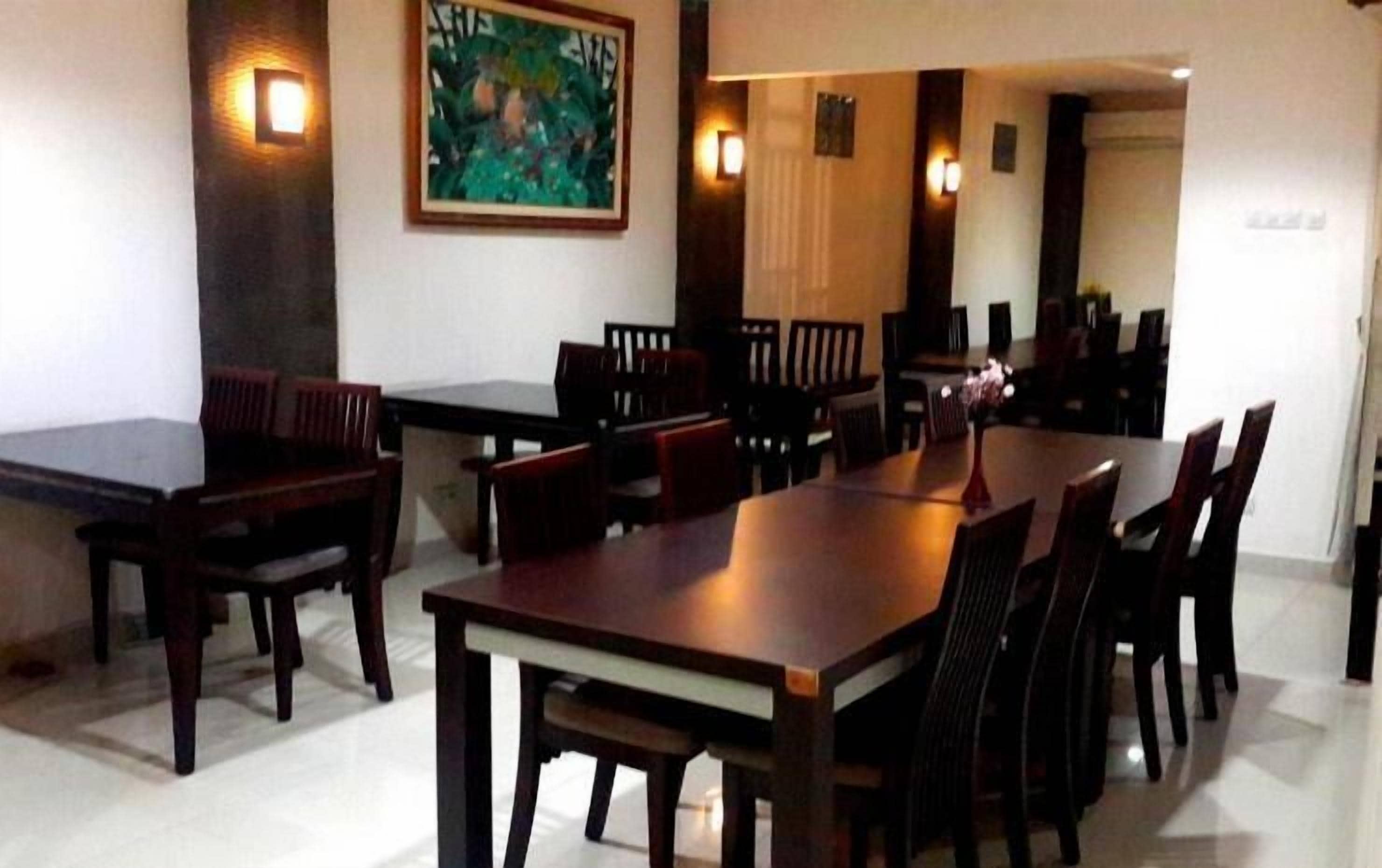 A dining area with multiple wooden tables and chairs, a painting on the wall, and wall-mounted lights.