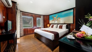A bedroom with a large bed, wooden walls, a painting of a beach scene, a TV, and a desk with a cup and fruit. - Huong Hai Sealife Cruise (Hong Gai)