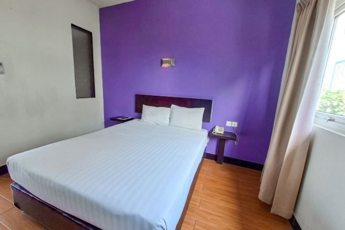 A hotel room with a large bed, a nightstand, a wall-mounted lamp, and a window with curtains.