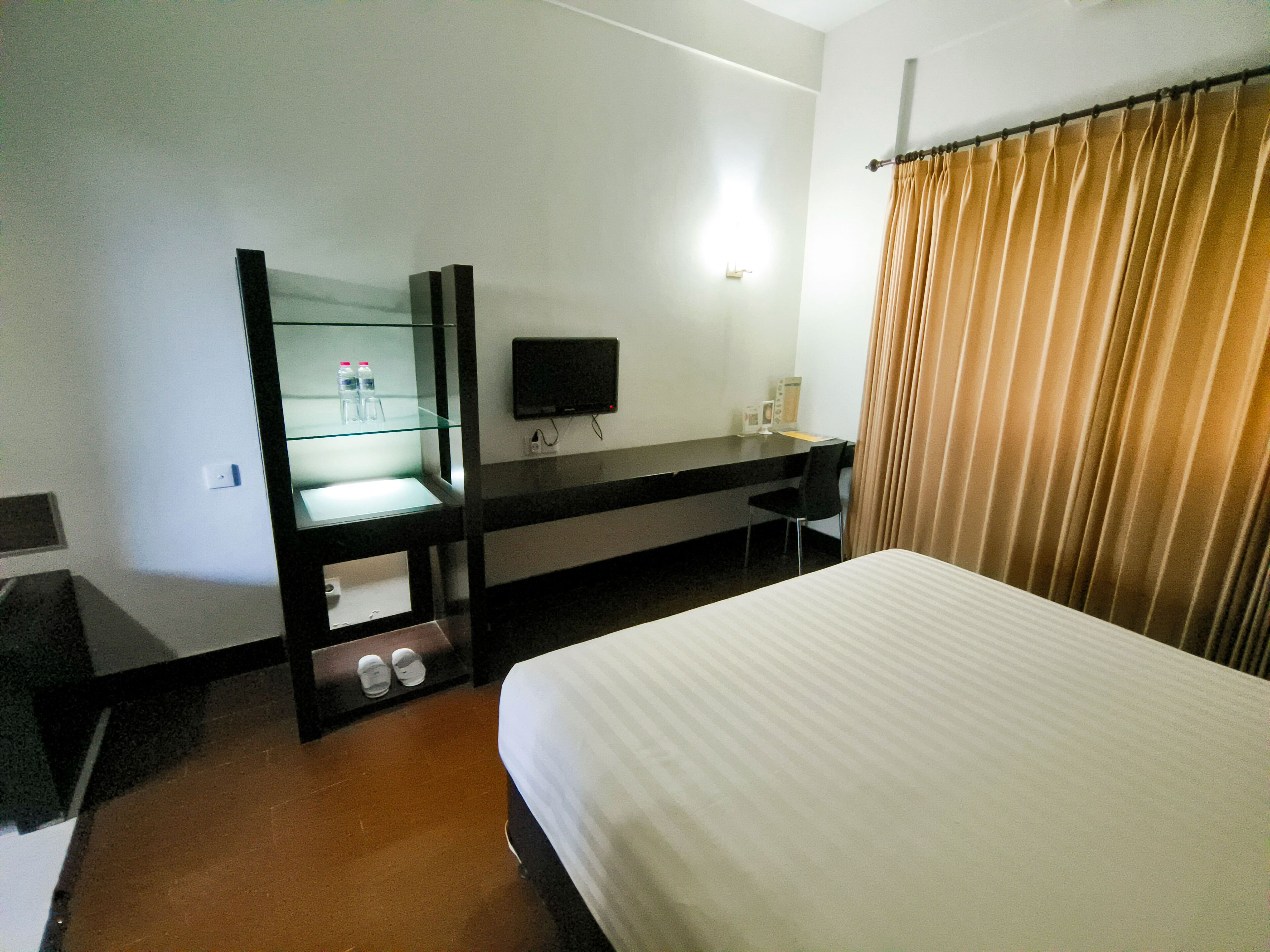 A hotel room with a bed, a desk with a flat-screen TV, a mirror, and a window with curtains.