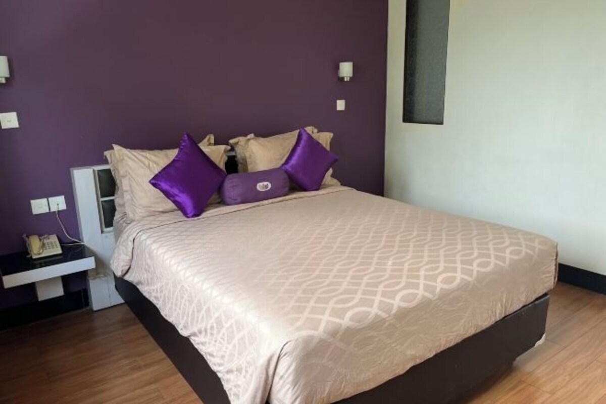 A neatly made bed with a patterned bedspread and multiple pillows, a bedside table with a lamp, and a wall-mounted light fixture.