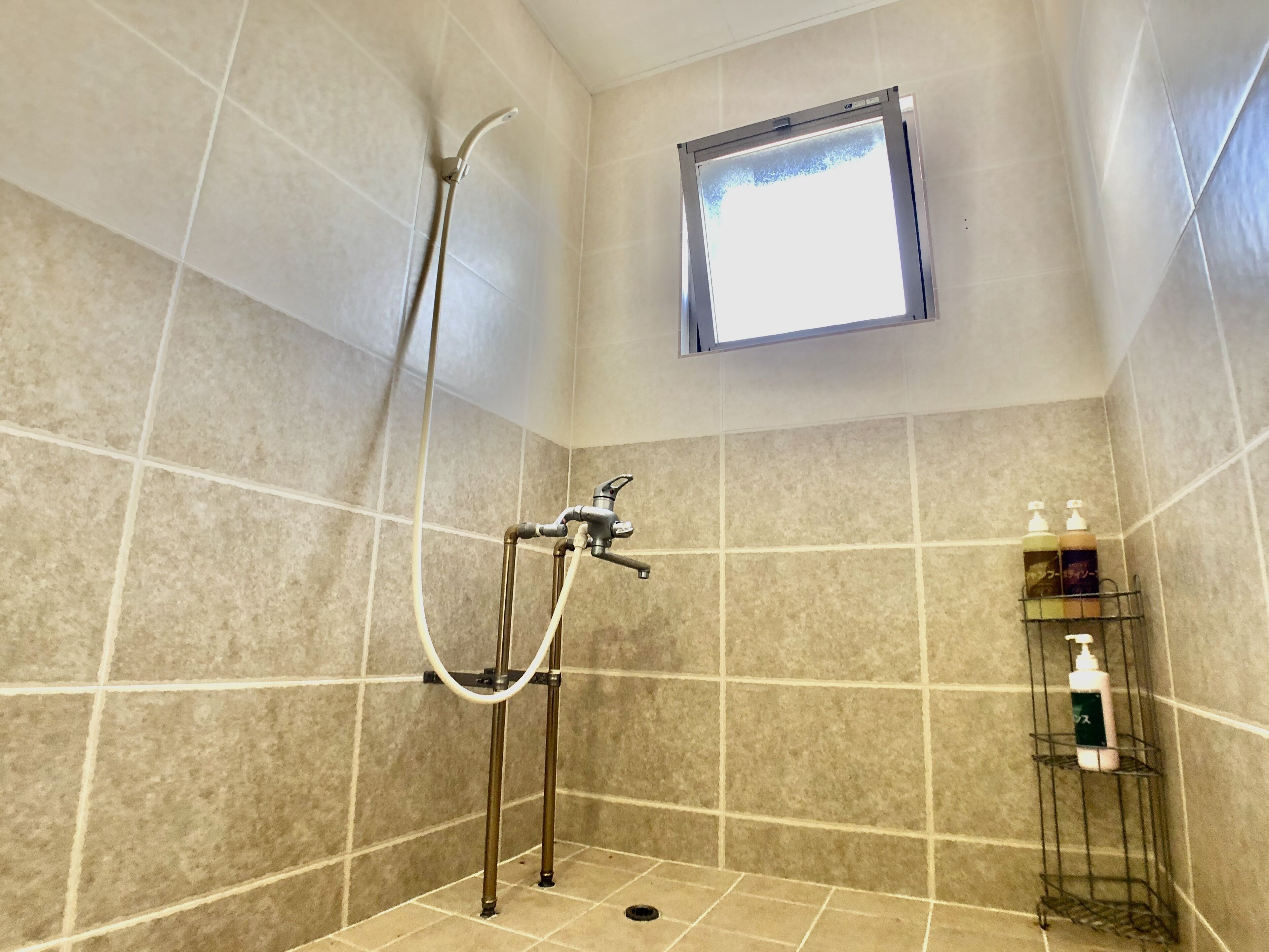 Pequeno espaço de chuveiro com azulejos, chuveiro manual, janela acima e prateleira com produtos de higiene pessoal.