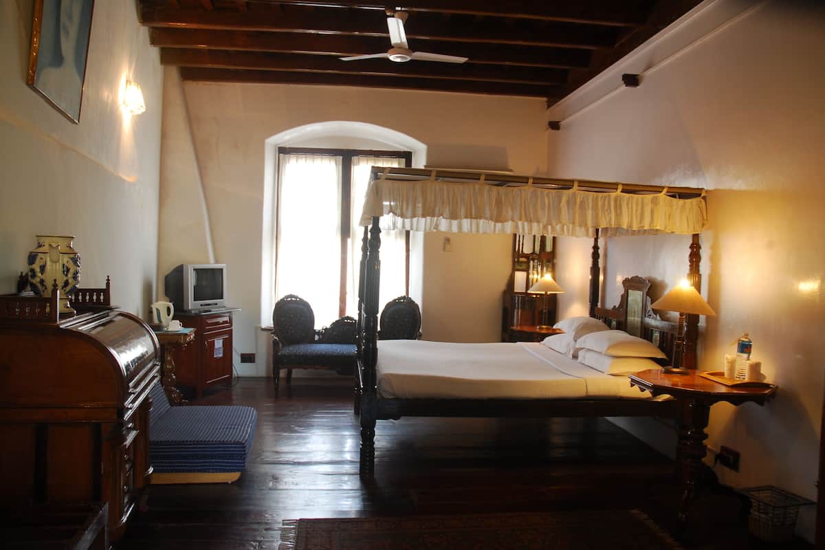 A traditional room with a four-poster bed, a desk with a chair, a television, and a large window with curtains.