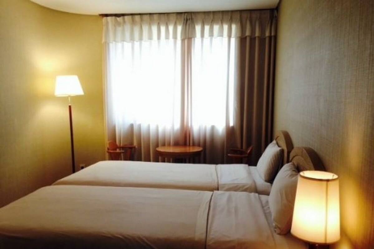 A hotel room with two beds, a small round table, a lamp, and a window with curtains.