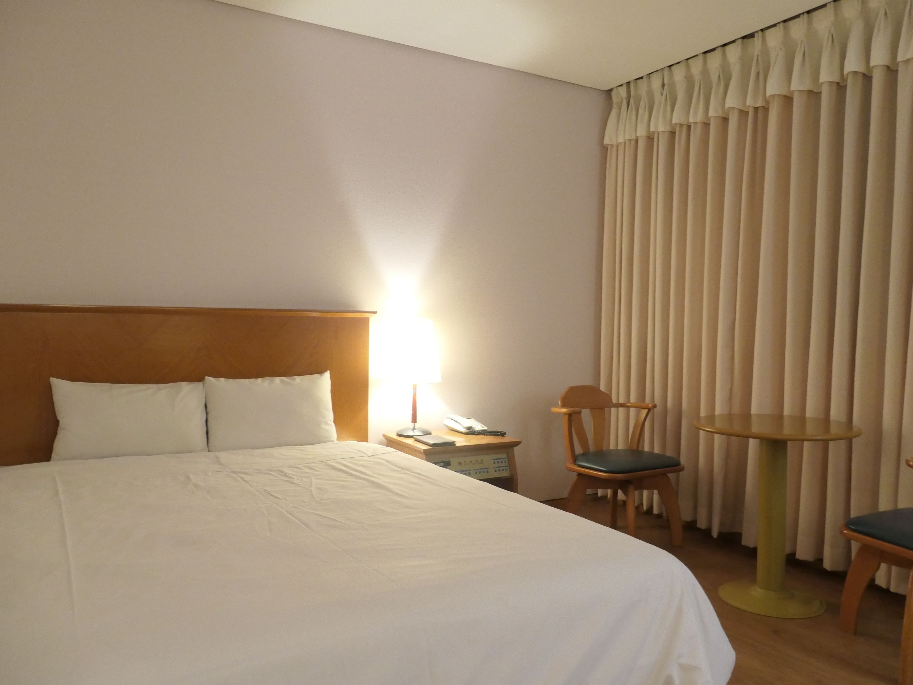 Una habitación de hotel con una cama grande, un cabecero de madera, una mesita de noche con lámpara, una silla y una pequeña mesa.