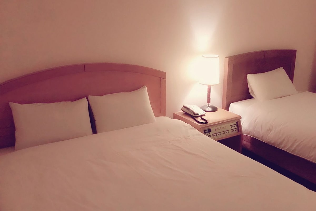 A hotel room with two beds, a bedside table with a lamp, and a telephone.
