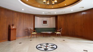 A conference room with a round table, two chairs, a green sofa, wooden panel walls, a chandelier, and a circular floor design. - Daedong Hotel (Jeju City)