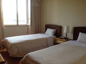 A hotel room with two beds, a nightstand with a lamp, and a window with curtains. - Daedong Hotel (Jeju City)