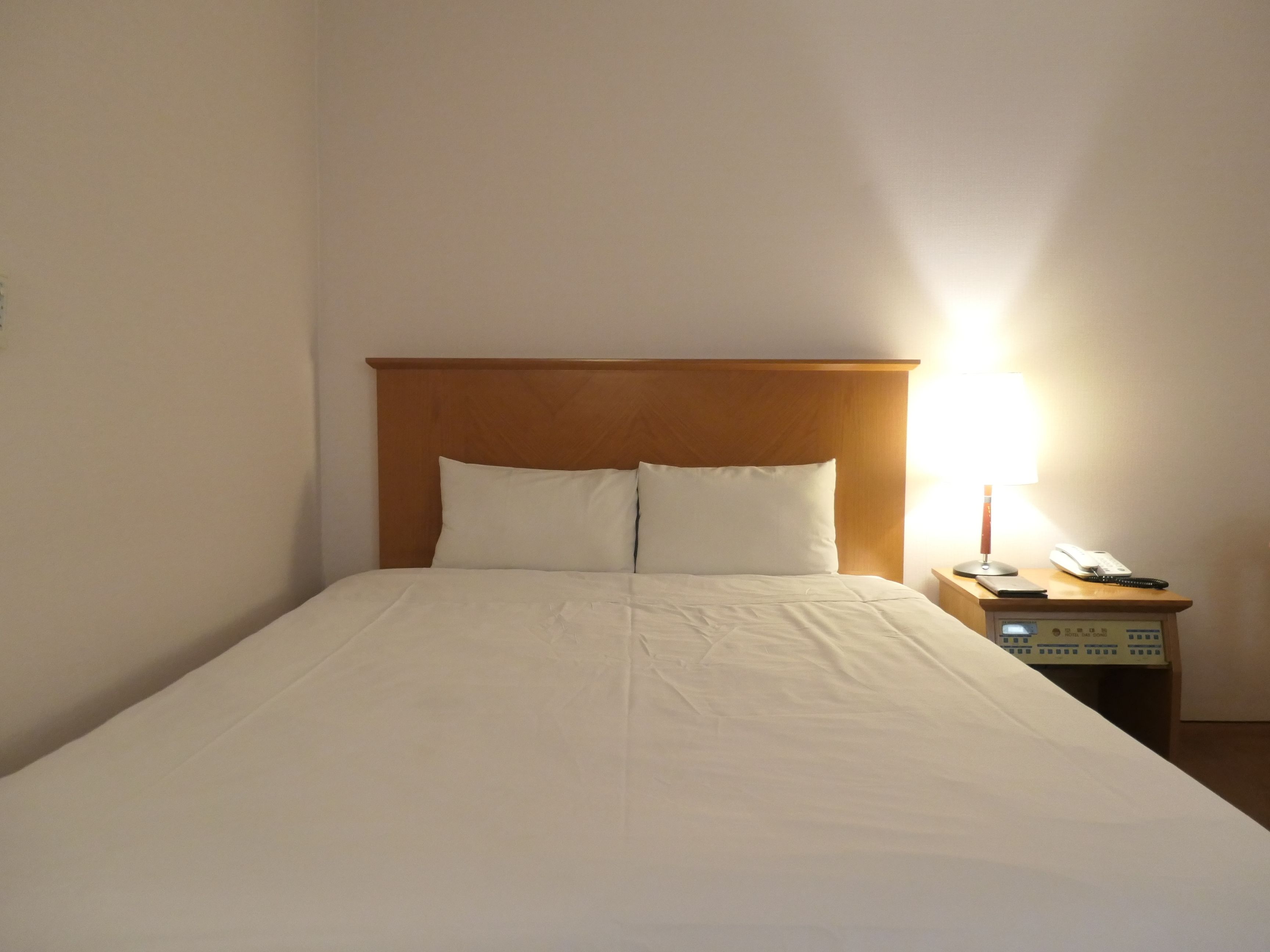 A single bed with a wooden headboard, two pillows, a bedside table with a lamp, and a phone.