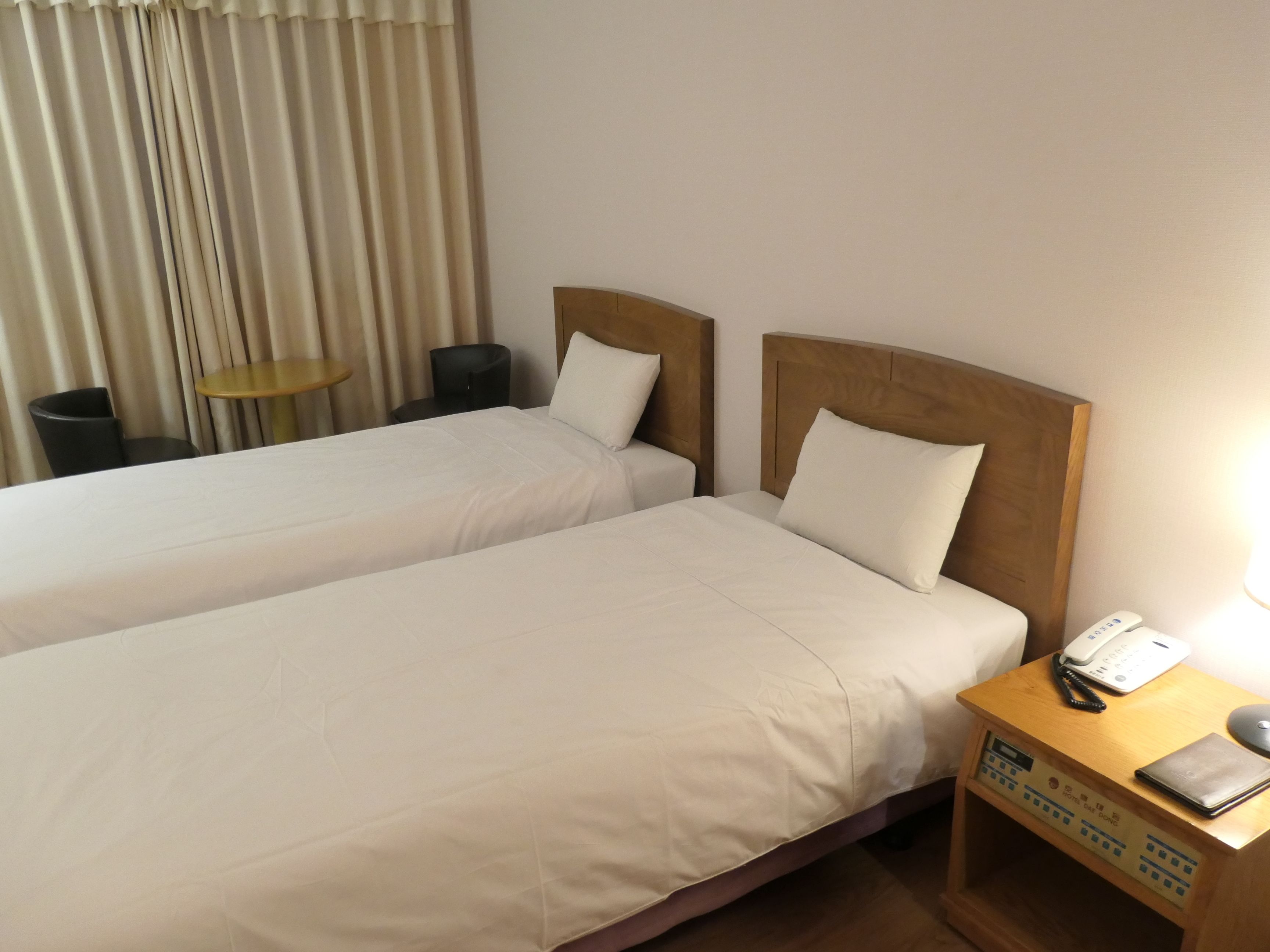 Habitación de hotel con dos camas, una mesita de noche con teléfono y lámpara, una pequeña mesa redonda y una silla.