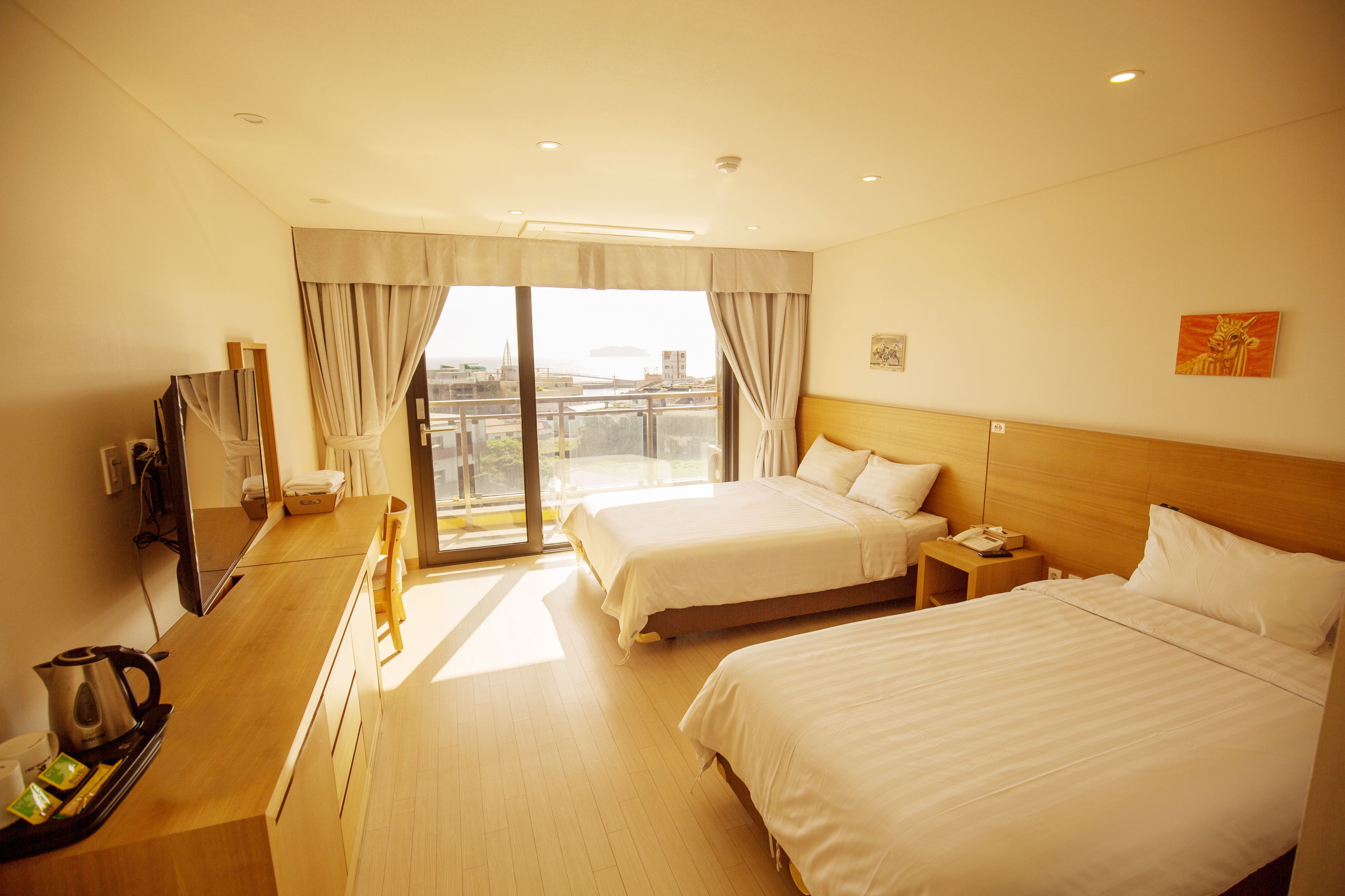 A hotel room with two beds, a desk with a kettle, a TV, and a large window with curtains.