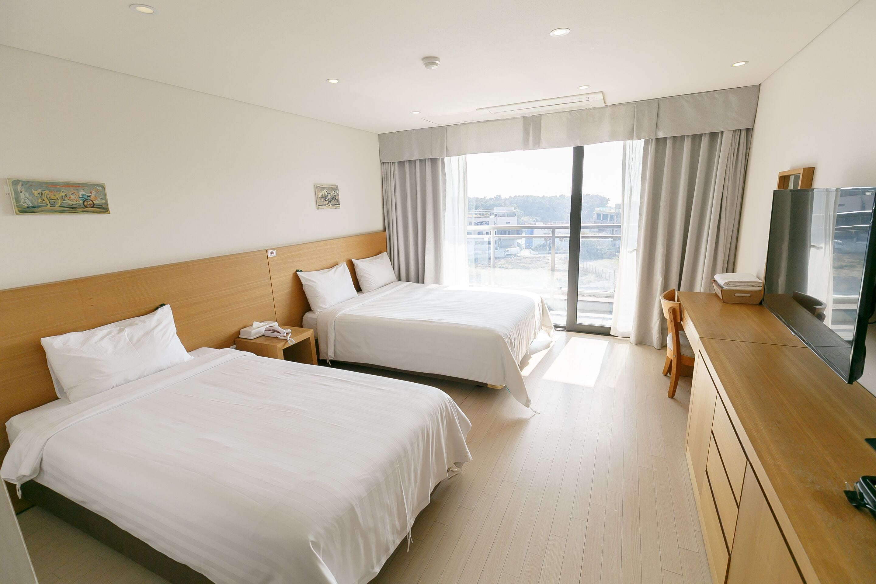 A hotel room with two beds, a wooden dresser, a television, and a large window with curtains.