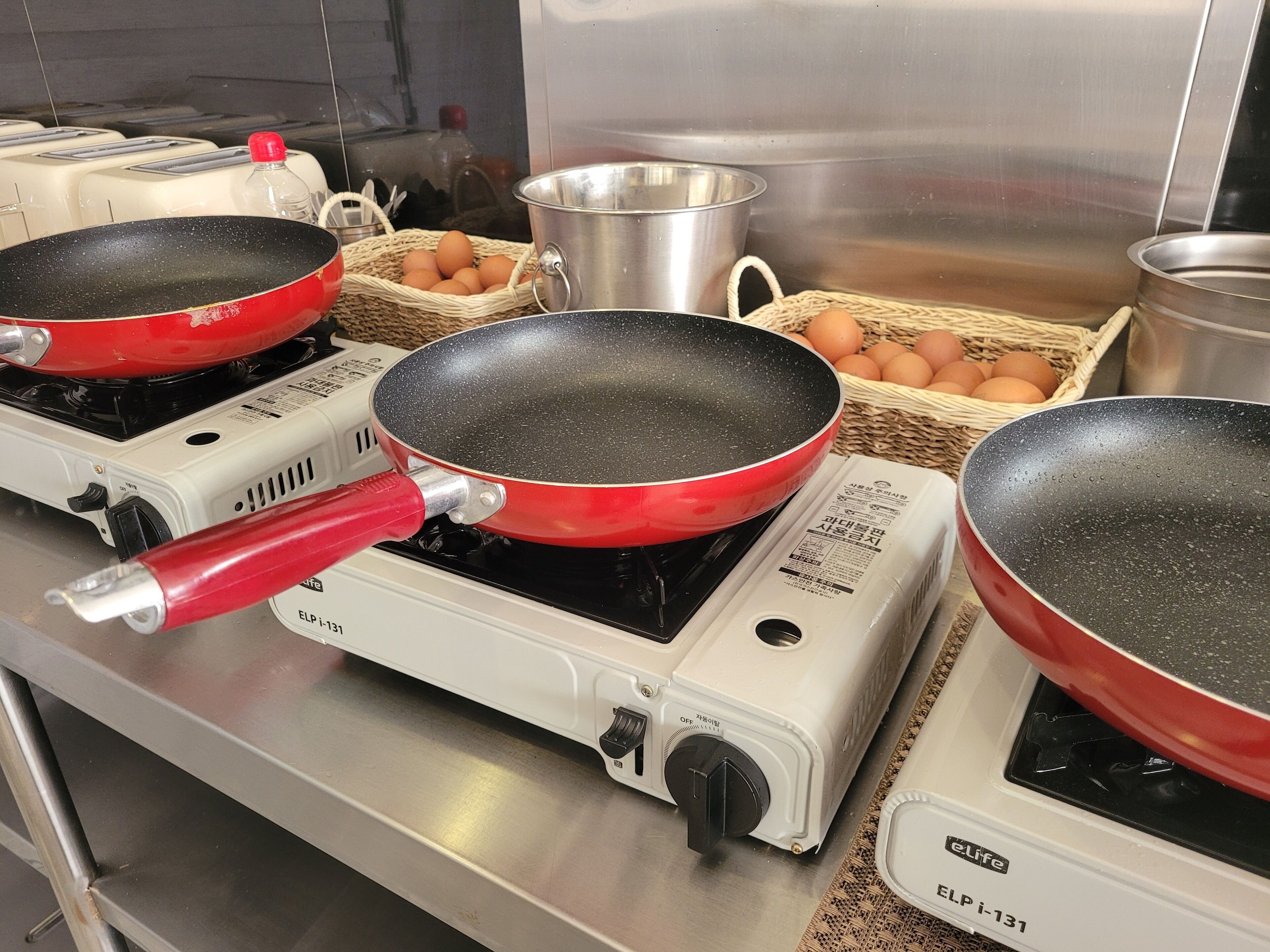 A kitchen with three electric fry pans on a stove, baskets of eggs, and stainless steel cookware.