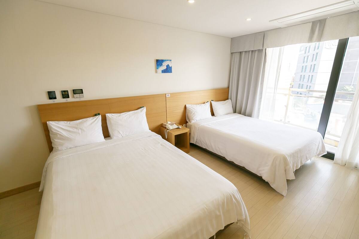 Hotellihuone, jossa on kaksi sänkyä, puinen pääty, pieni yöpöytä, jossa on puhelin, ja ikkuna, jossa on verhot.