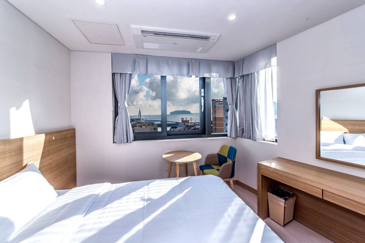 Hotellihuone, jossa on sänky, työpöytä, tuoli, pieni pöytä ja ikkuna, josta avautuu näkymä merelle ja rakennuksiin.
