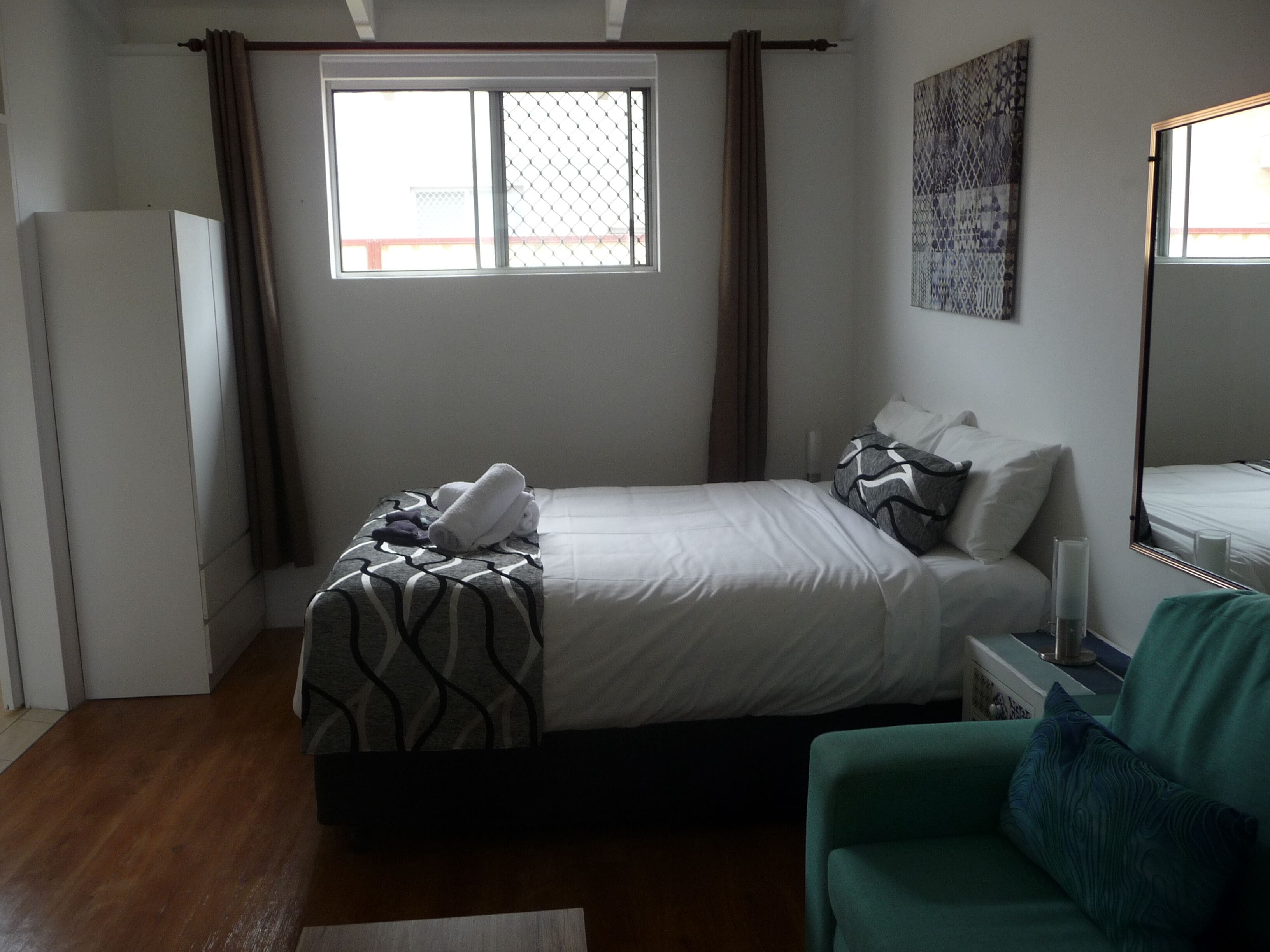 A bedroom with a bed, a green armchair, a window with curtains, a wall-mounted artwork, and a bedside table with a lamp.
