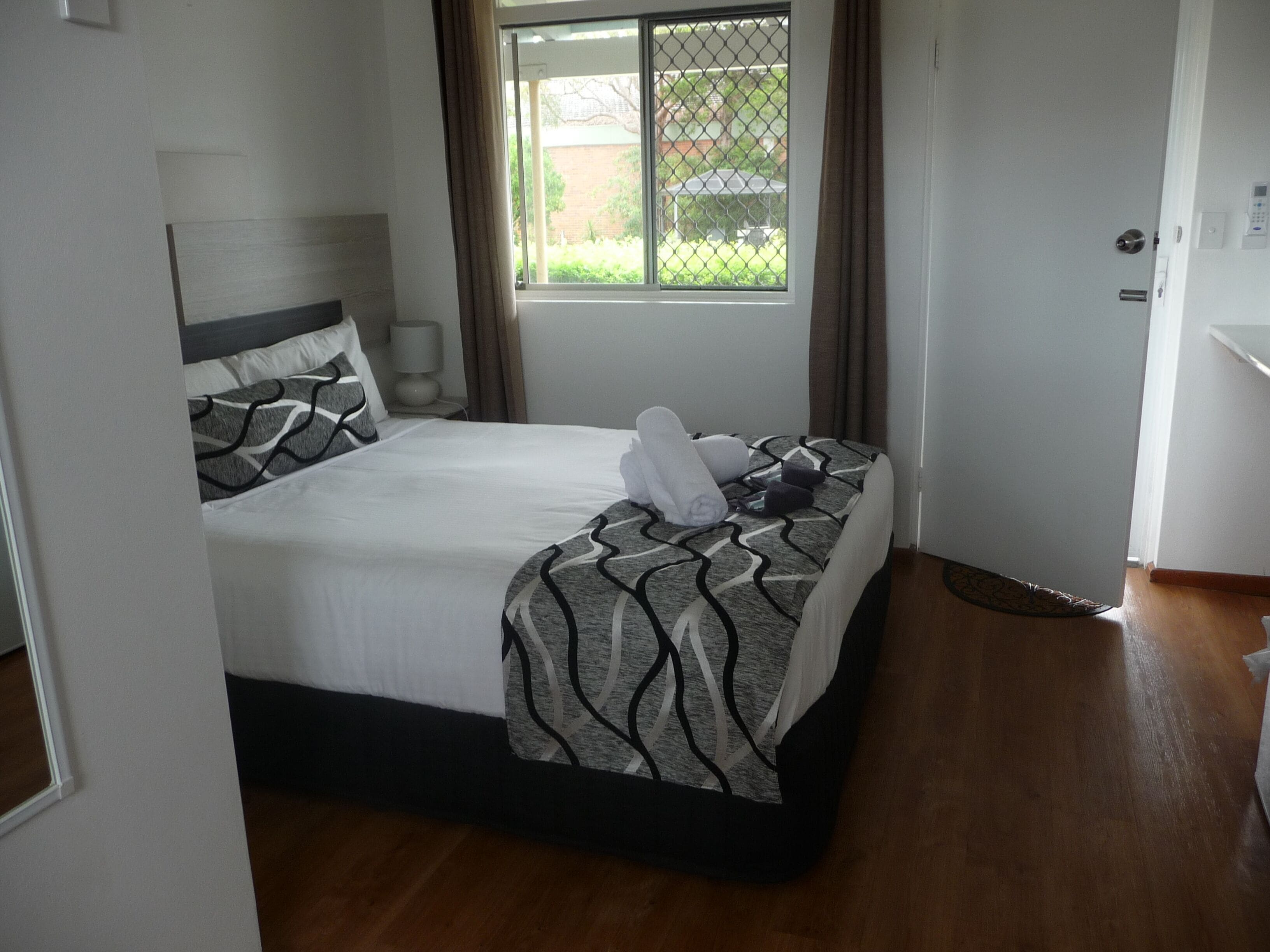 A neatly made bed with a patterned bedspread and pillows, a bedside lamp, and a view of a green outdoor area through a window.