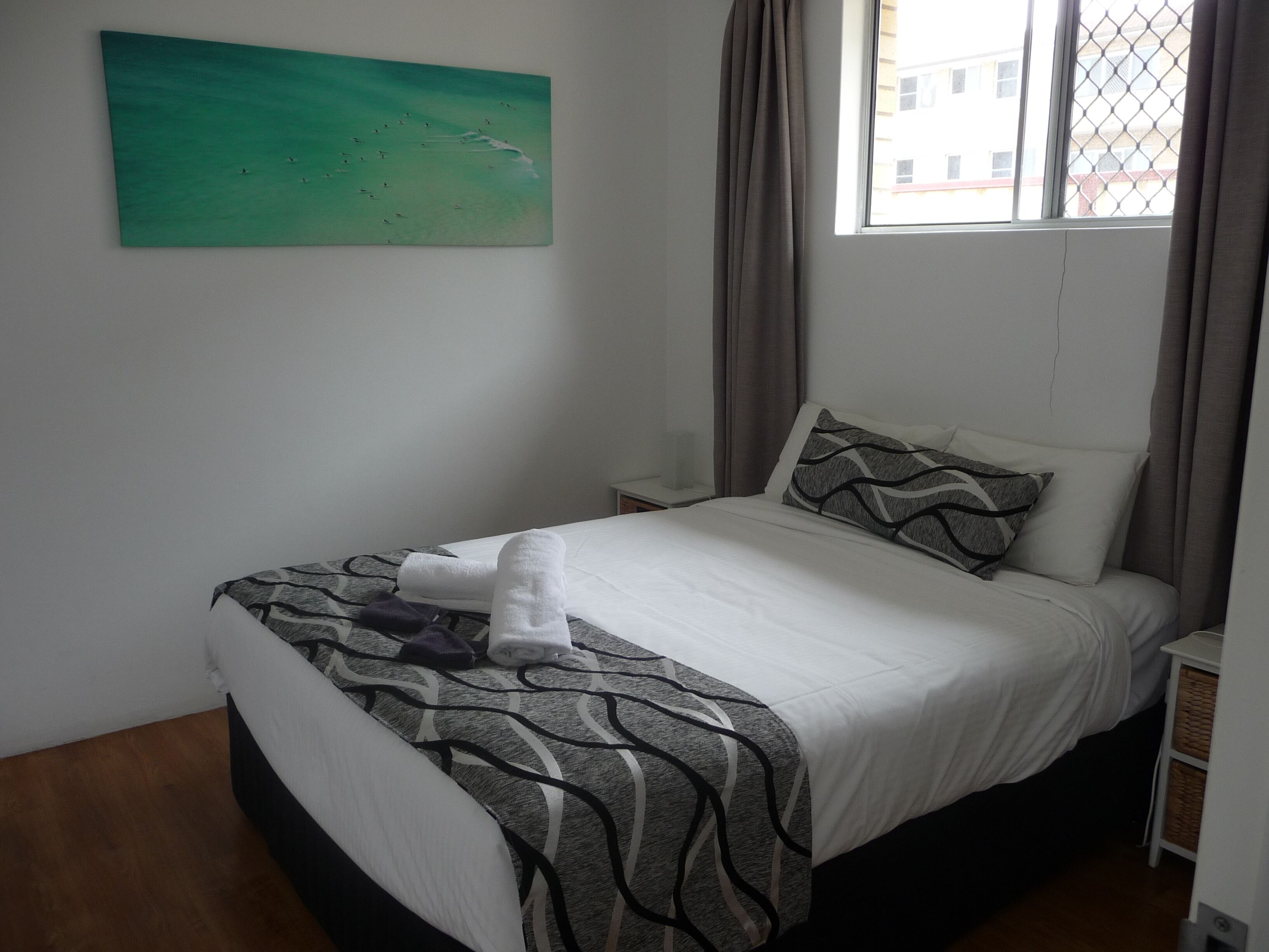 Un letto rifatto con cura, una coperta a fantasia, un cuscino con motivo ondulato e un asciugamano sul letto. Al muro, un quadro che raffigura un paesaggio, e una finestra con tende.