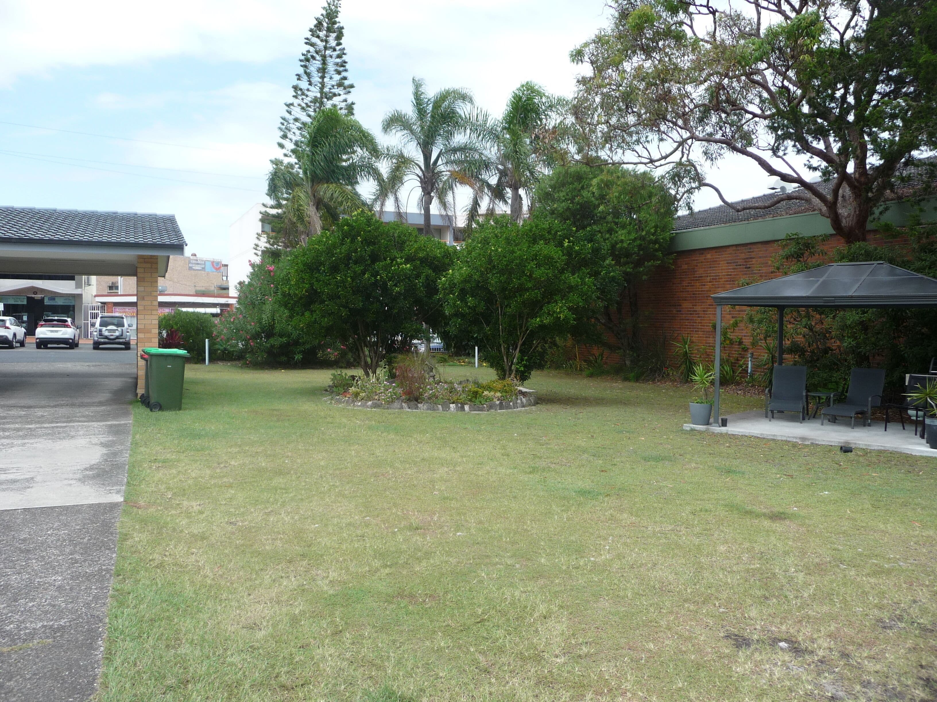 A parking lot with cars, a green trash bin, a grassy area, and a covered seating area with chairs and tables.