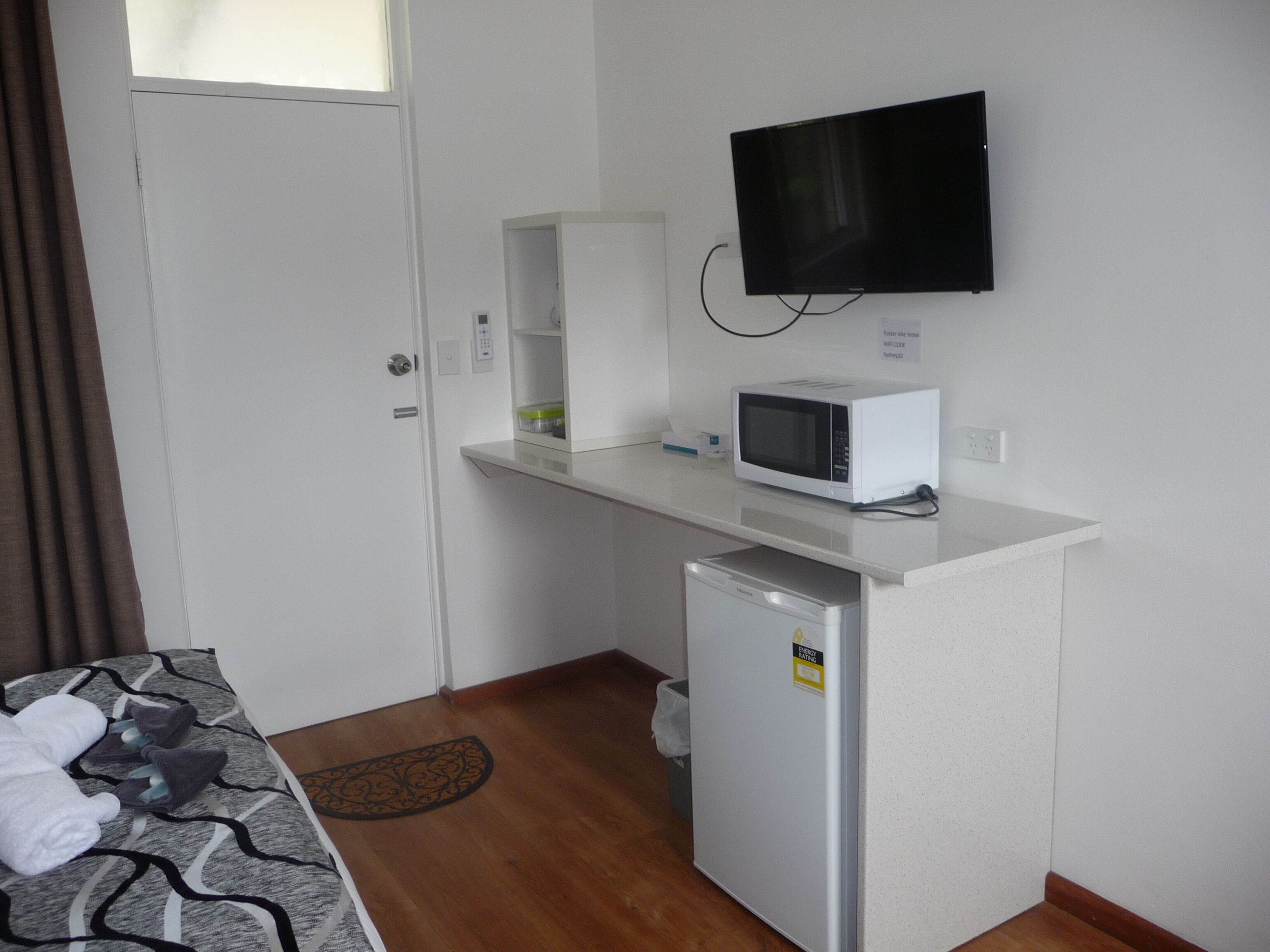 A small kitchenette with a microwave, a mini fridge, a TV mounted on the wall, and a bed with towels.