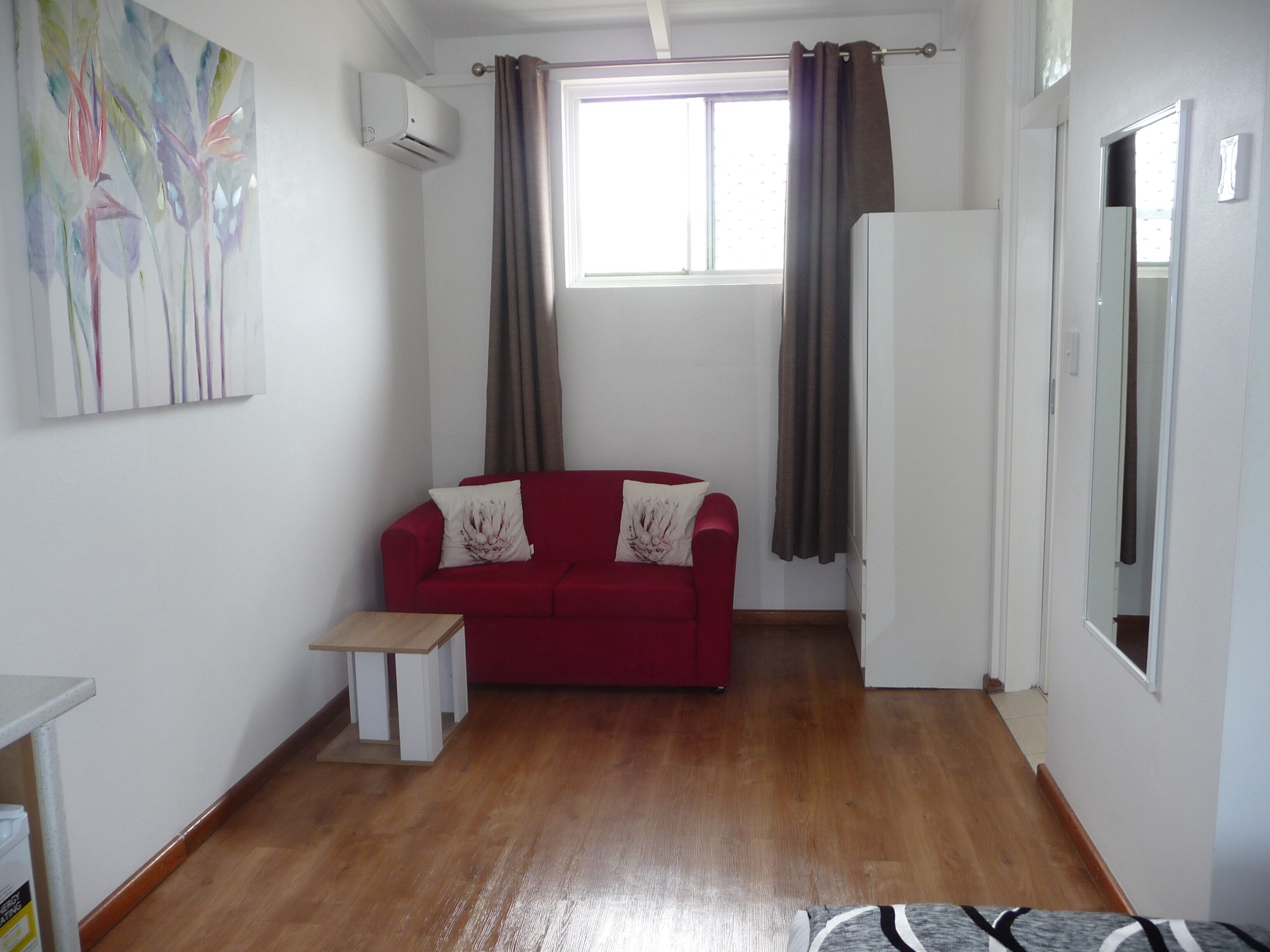 A room with a red sofa, a small white table, a window with curtains, and an air conditioning unit on the wall.