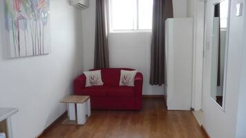 A room with a red sofa, a small white table, a window with curtains, and an air conditioning unit on the wall.