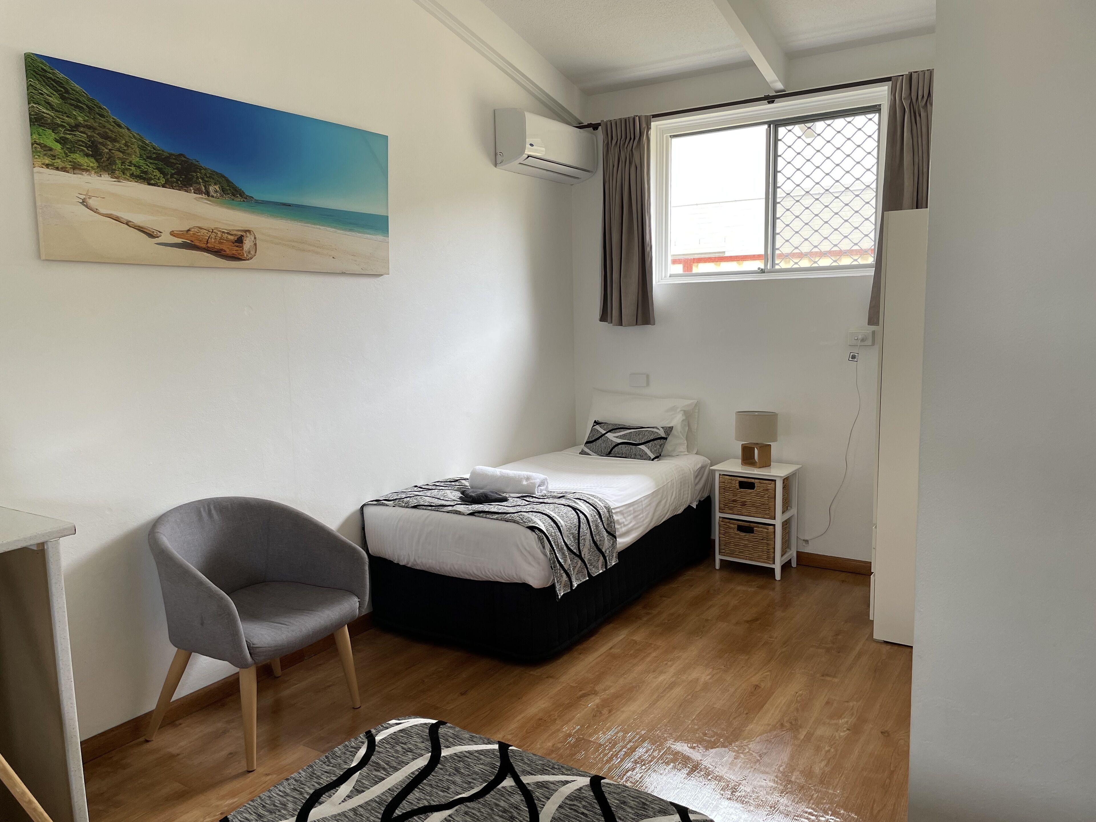Un dormitorio con cama, una silla, una mesita de noche con lámpara, un cuadro de una playa y un equipo de aire acondicionado.