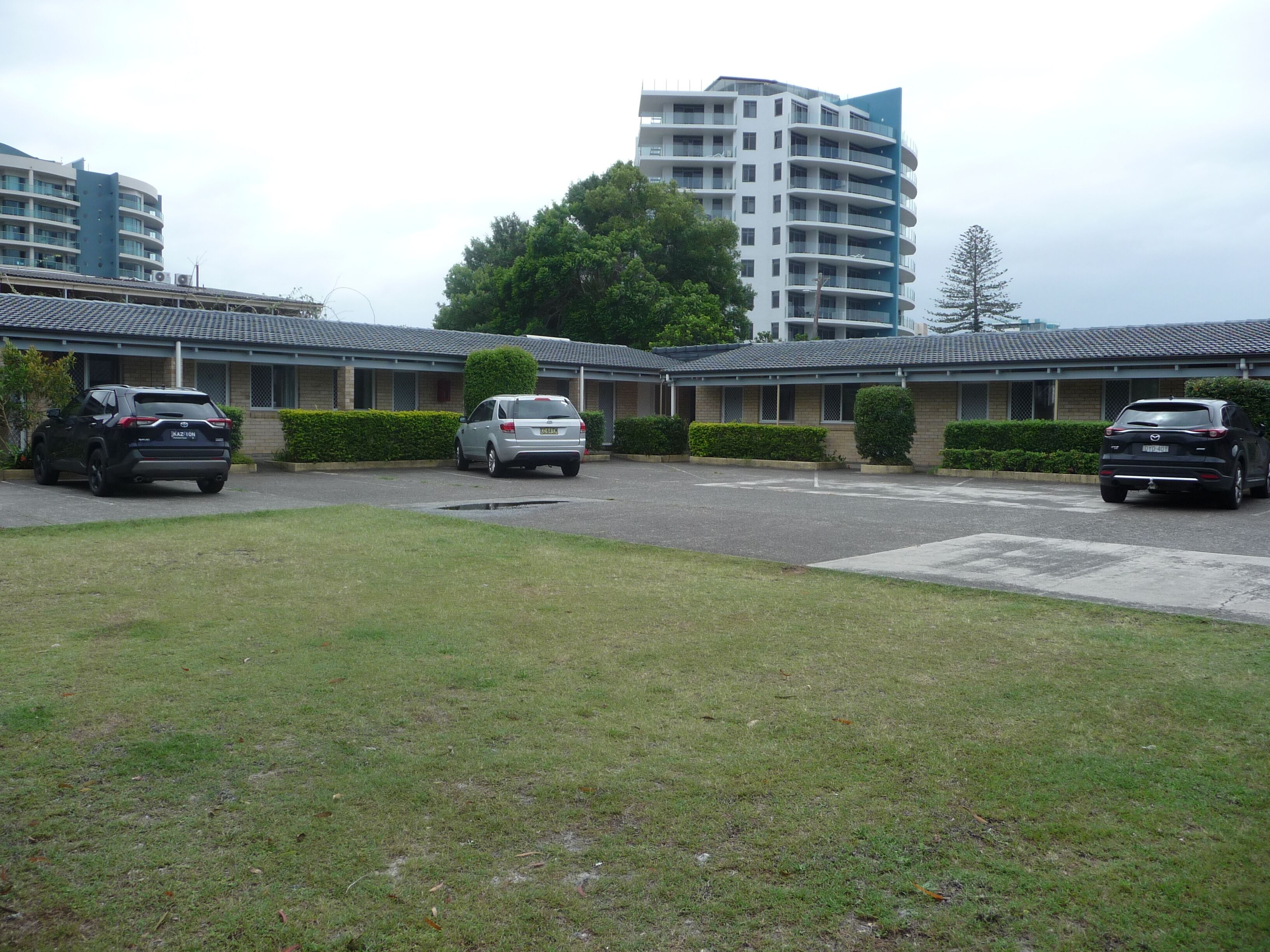 A parking lot with several cars, a building with a flat roof, and a tall residential building in the background.