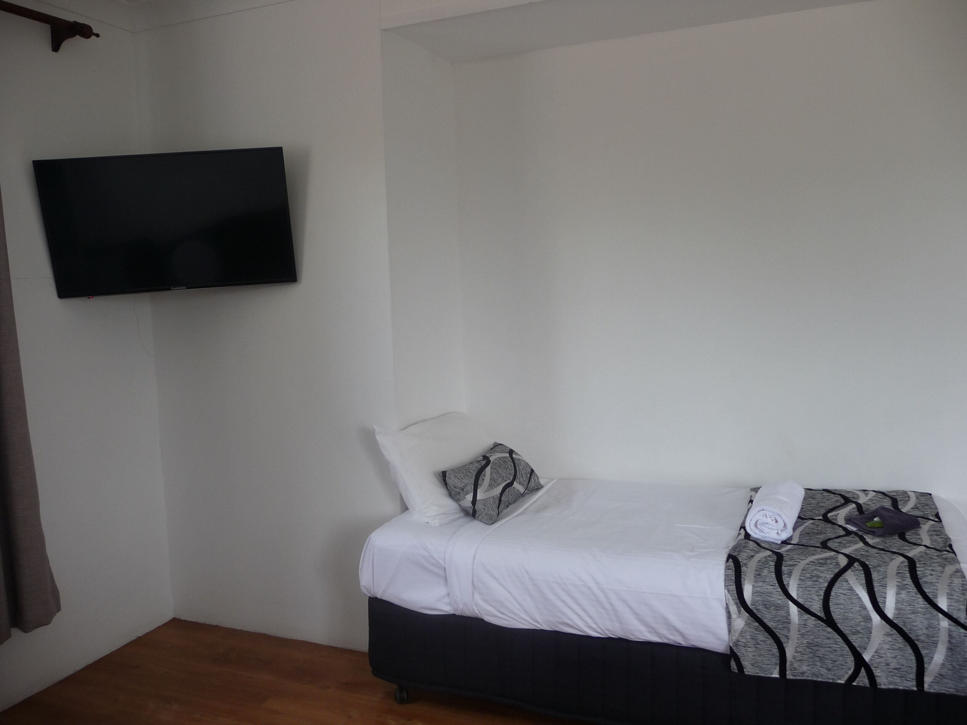 A single bed with a dark headboard, white bedding, and a patterned blanket. A flat-screen TV is mounted on the wall above the bed.