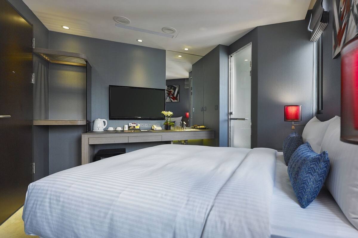 A modern hotel room with a large bed, a desk with a flat-screen TV, a coffee maker, and a nightstand with a lamp.