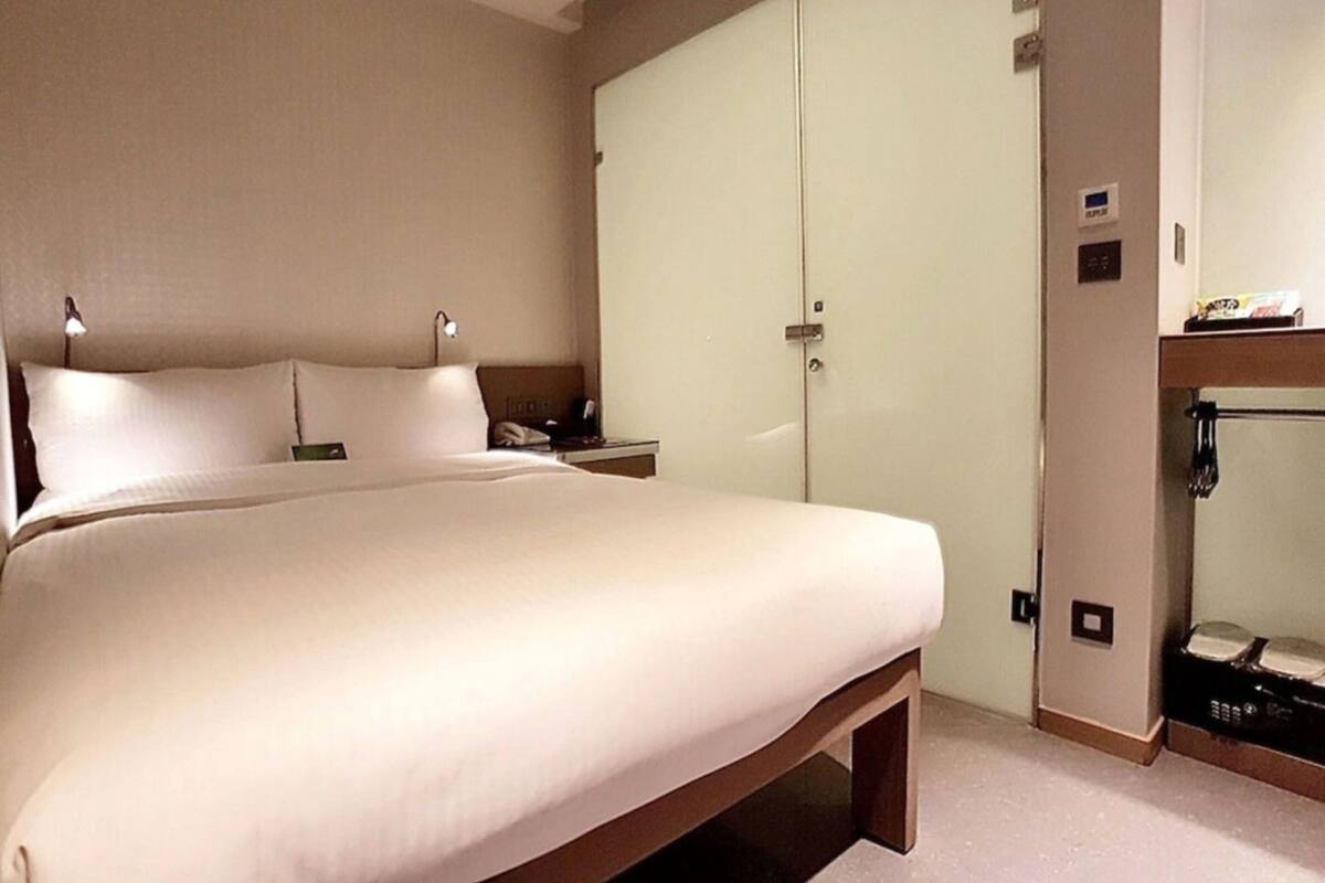 A hotel room with a large bed, a wardrobe, a bedside table, and a small shelf with items.