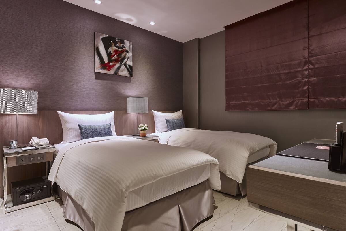 A hotel room with two beds, a bedside table with a lamp, a painting on the wall, and a large window with a blind.