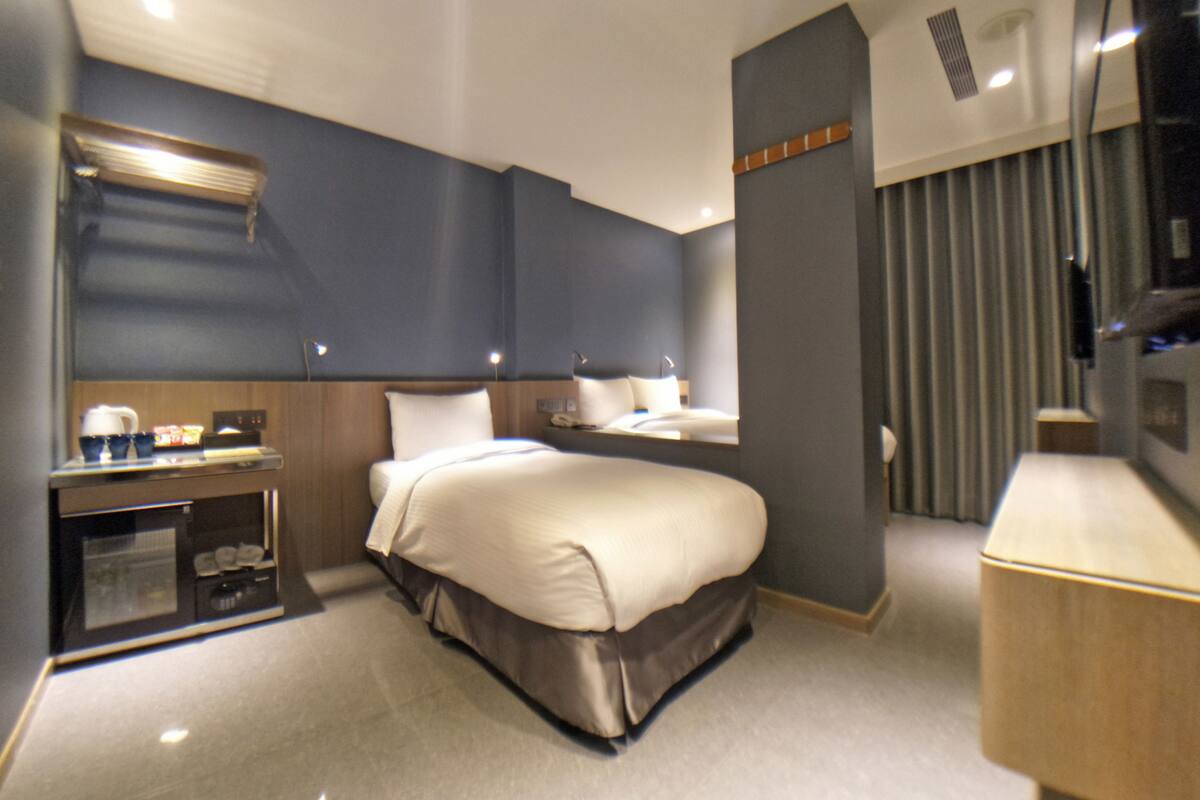 A hotel room with a bed, bedside table, lamp, and a small kitchenette area.