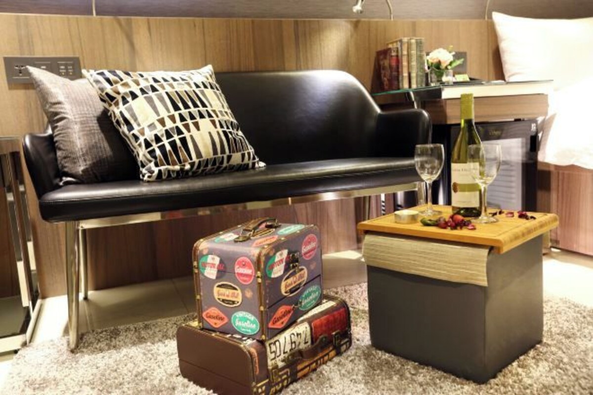 A modern living room with a black leather sofa, a patterned throw pillow, a small wooden coffee table with a bottle of wine and fruit, and a suitcase with stickers.