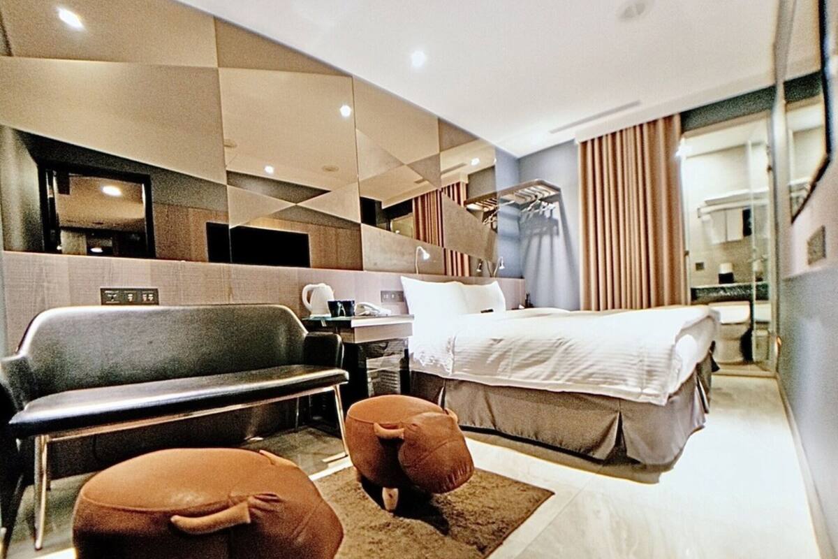 A modern hotel room with a bed, a sofa, two ottomans, a desk, and a mirror.