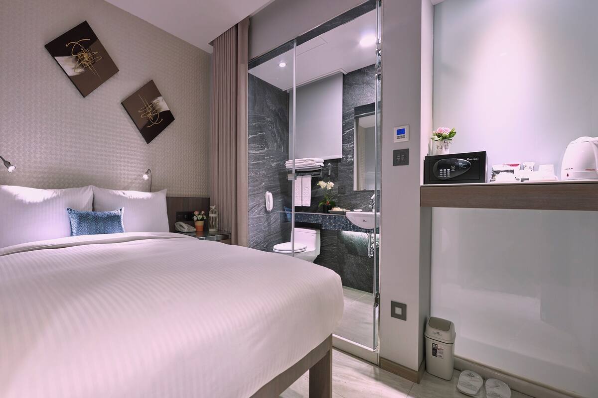 A modern hotel room with a large bed, a bedside table, a mirror, and a bathroom with a sink and toilet.